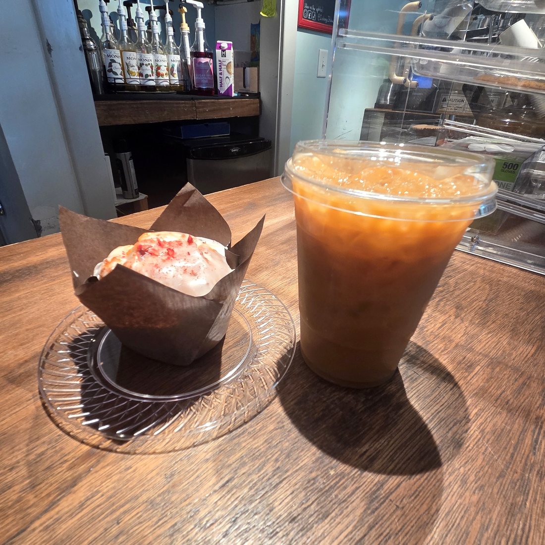 An iced coffee and a glazed muffin on the wooden counter at Icebergs, with the syrup rack and pastry case behind.